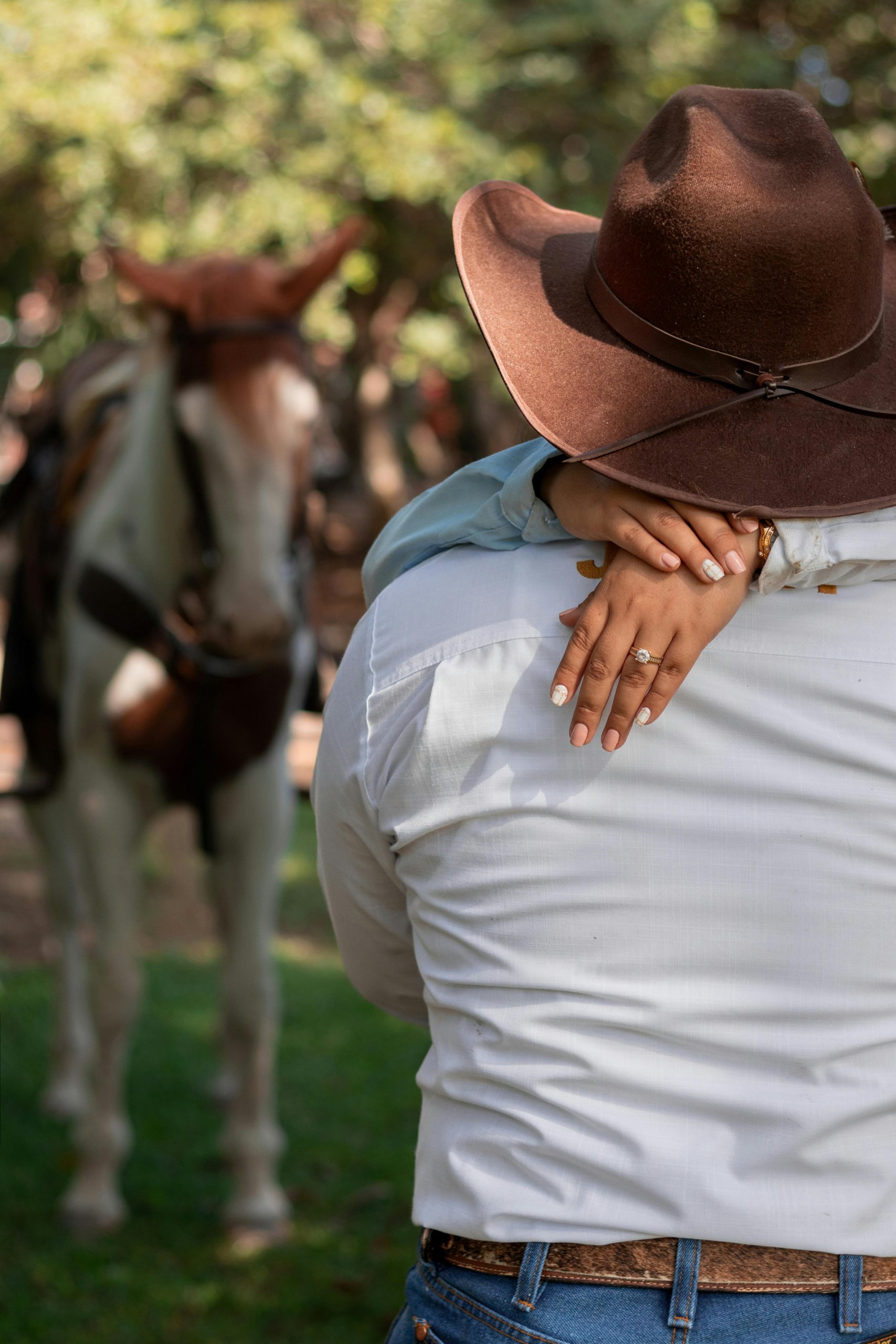 A couple in cowboy attire embraces in a pastoral setting with a horse nearby, conveying rural romance.